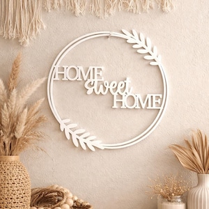 Op de afbeelding: Witte, cirkelvormige wanddecoratie met de woorden "HOME sweet HOME". Het ontwerp bevat bladeraccenten en wordt weergegeven tegen een neutraal getinte muur. Decoratieve elementen zoals vazen en een gevlochten mand zijn ook zichtbaar.