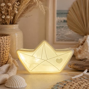 Può includere: Una luce notturna a forma di barchetta di carta luminosa. La luce bianco sporco presenta un dettaglio a cuore e linee geometriche. La lampada è su una superficie chiara, con conchiglie e un sottobicchiere intrecciato nelle vicinanze. Lo sfondo include piante secche e una scena di spiaggia incorniciata.