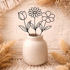 Könnte beinhalten: Eine kleine, gesprenkelte beige Vase hält drei schwarz gezeichnete Blumen: eine Tulpe, eine Gänseblümchen und eine fünfblättrige Blume. Die Vase steht auf einer strukturierten, cremefarbenen Oberfläche, mit getrockneten Gräsern und dekorativen Elementen im Hintergrund.