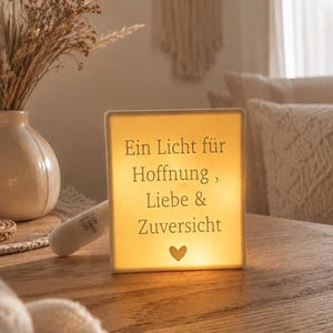 May include: A square, illuminated white light box featuring the German text 'Ein Licht für Hoffnung, Liebe & Zuversicht' and a small heart icon at the bottom. The device includes a white side switch with settings for on, off, and timer functions.