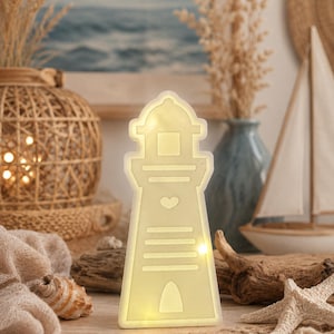 May include: A cream-colored, lighthouse-shaped LED light fixture featuring a cutout heart design, horizontal lines, and an arched base. The structure emits a soft, warm glow from within, highlighting its silhouette against the surrounding decor.
