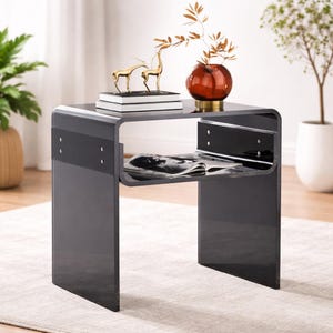 May include: A modern, glossy black side table with a curved design. It features a lower shelf holding a magazine and is decorated with a gold deer figurine, books, and a brown vase with decorative branches. The table is approximately 20 inches tall.
