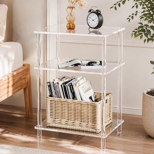 May include: A clear acrylic three-tier side table with a woven basket, books, a black alarm clock, and a decorative vase. The table is in a well-lit room with a wooden bed frame and a potted plant.