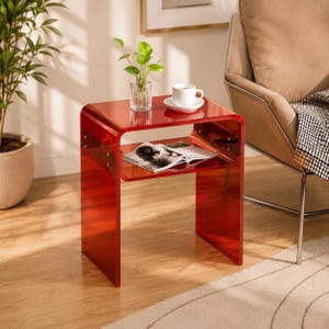 May include: A red translucent acrylic side table with a C-shaped design and a middle shelf. The table features visible metal hardware connecting the sides to the shelf. A small clear glass vase with a green plant and a white ceramic mug sit on top.