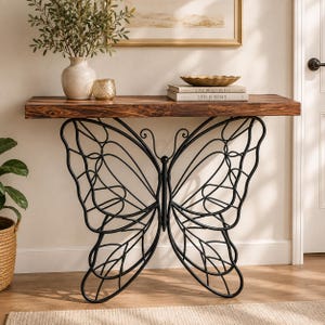 May include: A console table featuring a rectangular dark brown wood top and a black metal base shaped like a large butterfly. The table is positioned against a wall and supports a ceramic vase, a small gold candle holder, and two stacked books.