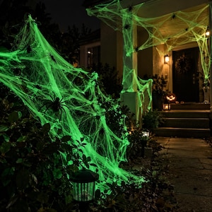 Pode incluir: Uma decoração de teia de aranha artificial verde neon brilhante drapeada sobre arbustos externos e a entrada de uma varanda. A teia apresenta várias figuras de aranha de plástico preto presas aos fios para uma decoração de Halloween.