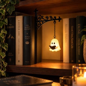 Puede incluir: Un soporte de pared de metal negro con un diseño de volutas ornamentadas sostiene una pequeña figura de fantasma blanco brillante suspendida por una cadena. El fantasma tiene forma de sábana con dos ojos negros y sostiene un libro negro abierto.