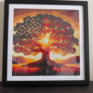 Puede incluir: Obra de arte enmarcada que presenta un diseño de árbol con un fondo de puesta de sol. El follaje del árbol incorpora un patrón de la bandera estadounidense. La obra está compuesta por pequeños elementos coloridos, creando un efecto de mosaico. El marco es negro y la obra se muestra sobre un paspartú blanco.