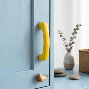 Pode incluir: Um puxador de armário curvo amarelo brilhante montado numa porta de armário de madeira azul-clara. Abaixo do puxador, encontra-se um pequeno botão de madeira esculpida em forma de pássaro. A porta apresenta um veio de madeira visível.