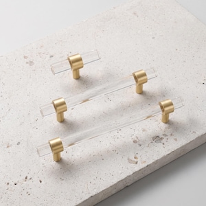May include: Three cabinet pulls featuring clear, cylindrical acrylic bars accented with brushed gold metal mounting posts. The set includes one small knob-style pull and two longer, horizontal bar-style pulls, each with two gold metal supports.