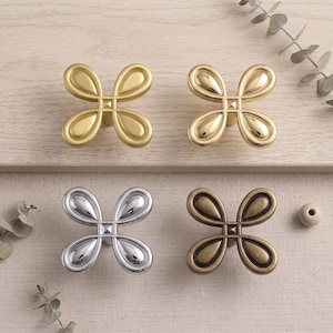 Könnte beinhalten: Vier Schrankknöpfe aus Metall in Form von vierblättrigen Blumen. Der Knopf oben links hat ein mattes Gold-Finish, oben rechts ein poliertes Gold-Finish, unten links ein poliertes Chrom-Finish und unten rechts ein antikes Messing-Finish.