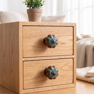 May include: A light-colored wooden storage unit with two drawers, each featuring a round, fluted knob with a metallic bronze center and a teal-colored glazed finish. The wood grain is visible on the drawer fronts and the frame of the unit.