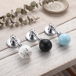 May include: Three drop-style cabinet pulls with polished chrome circular bases and hinged arms. The first pull features a white sphere with black paint splatters, the second a black marble-patterned sphere, and the third a solid light blue sphere.
