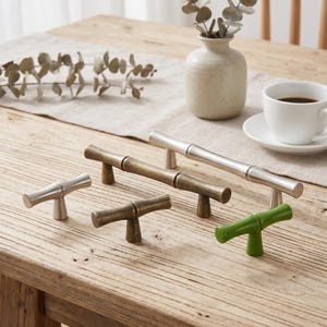May include: A collection of five cabinet pulls and knobs featuring a bamboo-inspired design. The set includes two silver-toned metal pieces, two antique brass-toned metal pieces, and one glossy green-finished piece, all with a T-shaped structure.