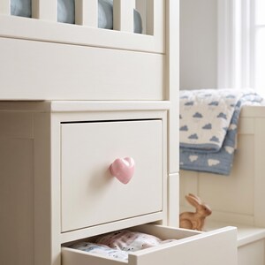 Puede incluir: Un mueble de almacenamiento de madera color crema con un cajón que tiene un pomo brillante en forma de corazón de color rosa claro. Debajo del cajón hay un compartimento abierto con tela estampada doblada. El mueble tiene un acabado mate y liso.