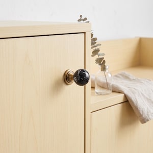 Könnte beinhalten: Ein runder Möbelknopf mit einer schwarzen, marmorierten Kugel mit weißen Adern, montiert auf einem kreisförmigen Sockel aus antikem Messing. Der Knopf ist an einer hellen Holzschranktür mit glatter, natürlicher Maserung befestigt.