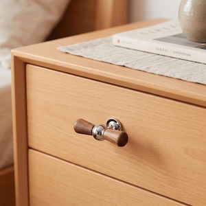 May include: A drawer pull featuring a horizontal wooden handle with a smooth, tapered shape, connected to a central polished chrome sphere and a circular chrome base plate, mounted on a light-toned wooden drawer front with a visible grain.