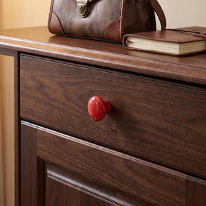 May include: A glossy, round, bright red cabinet knob is attached to the center of a dark brown wooden drawer front. The drawer is part of a wooden furniture piece with a visible grain pattern, featuring a brown leather bag and a notebook on top.