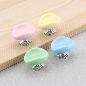 Pode incluir: Quatro puxadores de armário com topos lisos em forma de seixo nas cores rosa pastel, amarelo pálido, verde claro e azul celeste. Cada puxador possui uma base de metal prateado polido e um acabamento brilhante, com um design simples.