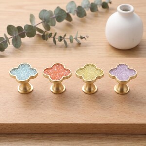 May include: Four clover-shaped cabinet knobs with a brushed gold metal finish and inlaid textured centers in light blue, orange, yellow, and purple. Each knob features a four-leaf clover silhouette with a central pedestal base for mounting.