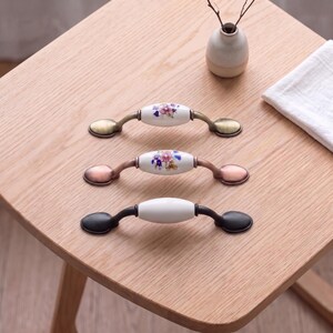 May include: Three cabinet pulls with oval ceramic centers and metal ends. The top pull has a brass finish and floral pattern, the middle has a copper finish and floral pattern, and the bottom pull has a matte black finish with a plain white center.