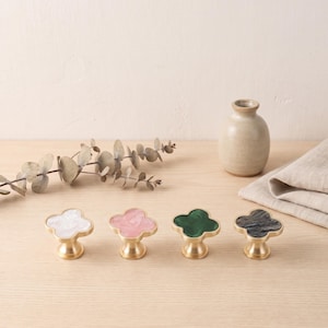 May include: Four clover-shaped cabinet knobs with brass-toned metal bases. Each knob features a distinct inlaid top: one white marbled, one light pink marbled, one solid dark green, and one dark grey marbled finish, arranged in a horizontal row.