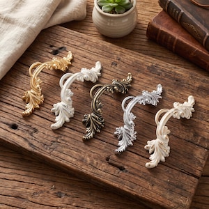 May include: Five curved drawer pulls featuring ornate leaf and scroll detailing. The set includes one gold, one white, one antique bronze, one silver, and one white with gold-toned highlights, all arranged in a row on a rustic wooden surface.