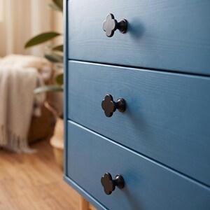 May include: A blue wooden dresser featuring three horizontal drawers, each equipped with a matte black clover-shaped knob. The furniture piece displays a visible wood grain texture across its front surface, with the drawers stacked vertically.