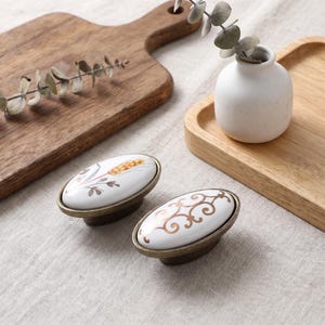 Può includere: Due pomelli per armadietto di forma ovale con superficie in ceramica bianca e base in metallo color bronzo. Un pomello presenta un disegno dipinto di spiga di grano, mentre l'altro mostra un motivo a filigrana dorata a spirale.