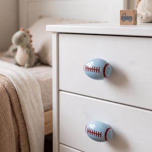 May include: Two light blue drawer knobs shaped like American footballs, featuring dark red laces and stripes. These oval-shaped ceramic knobs are attached to a white wooden dresser, providing a functional and thematic accent to the furniture piece.