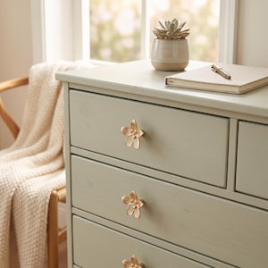 May include: A light sage green wooden dresser featuring three gold-toned metal drawer pulls shaped like five-petaled flowers. The dresser top holds a small potted succulent in a beige ceramic pot and a closed notebook with a pen resting on top.