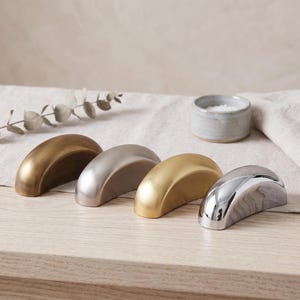 May include: Four semi-circular metal drawer pulls arranged in a row. The finishes include a dark antique brass, a brushed nickel, a satin gold, and a polished chrome. Each handle features a smooth, curved arch design with a solid, heavy appearance.