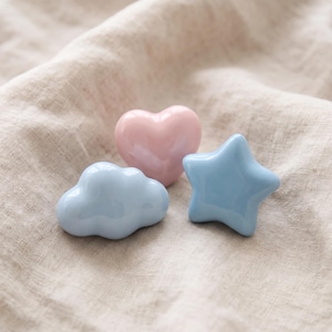 May include: Three ceramic drawer knobs with a smooth, glossy finish. One knob is shaped like a light pink heart, one is a light blue cloud, and the third is a light blue five-pointed star. Each piece features a rounded, puffed design.