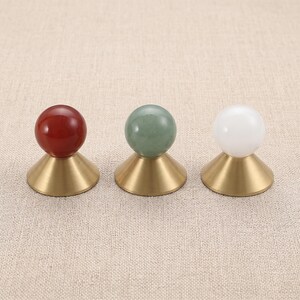 Op de afbeelding: Drie decoratieve knoppen met bolvormige bovenkanten in rood, groen en wit, elk gemonteerd op een conische basis van geborsteld messing. De set bevat een dieprode bol, een gedempte groene bol en een ondoorzichtige witte bol met metalen afwerking.