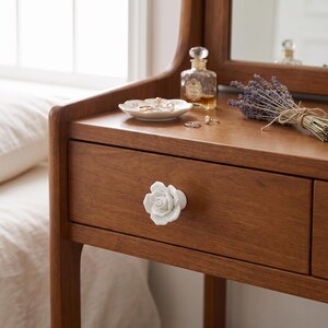 May include: A white ceramic drawer knob shaped like a blooming rose attached to a wooden vanity table. The vanity surface holds a small white scalloped dish containing rings, a glass perfume bottle, and a bundle of dried lavender stems.