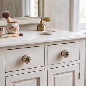 May include: A white wooden cabinet featuring two drawers with round, fluted ceramic knobs. Each knob is decorated with a hand-painted orange floral pattern and green leaves, finished with a central antique brass-toned metal accent piece.