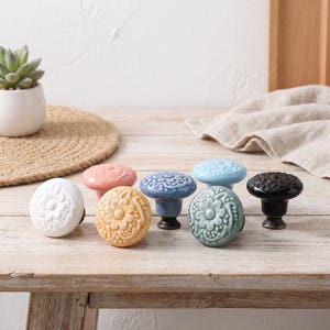 Peut inclure: Un ensemble de six boutons de placard ronds en céramique de couleurs blanche, rose, jaune, bleue, sarcelle et noire. Chaque bouton présente un motif floral en relief sur le dessus et une base en métal sombre pour les portes de meubles.