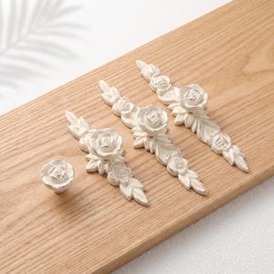 May include: A collection of four off-white cabinet hardware pieces featuring sculpted rose and leaf designs with subtle gold-toned highlights. The set includes one round knob and three elongated drawer pulls, each displaying detailed floral relief patterns.