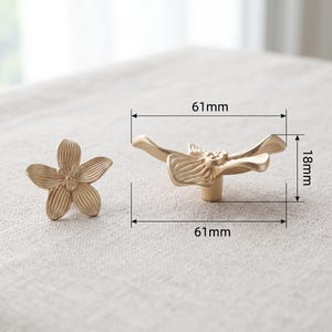 K&ouml;nnte beinhalten: Zwei goldfarbene Schrankkn&ouml;pfe aus Metall in Form von f&uuml;nfbl&auml;ttrigen Blumen. Ein Knopf ist von vorne abgebildet, der andere von der Seite, mit technischen Linien, die eine Breite von 61 mm und eine H&ouml;he von 18 mm f&uuml;r den Beschlag angeben.