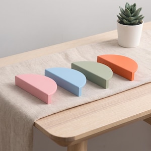 Puede incluir: Cuatro tiradores de mueble semicirculares en colores pastel mate, específicamente rosa, azul claro, verde salvia y naranja. Cada tirador presenta un borde superior plano y una cara frontal curva, diseñado para montarse en puertas o cajones.