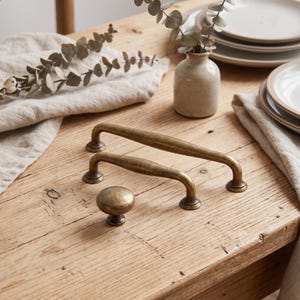 Peut inclure: Deux poignées de tiroir allongées en métal et un bouton de placard rond, tous dotés d'une finition en laiton antique patiné avec une surface texturée. Les pièces de quincaillerie sont disposées ensemble, affichant une teinte métallique uniforme.