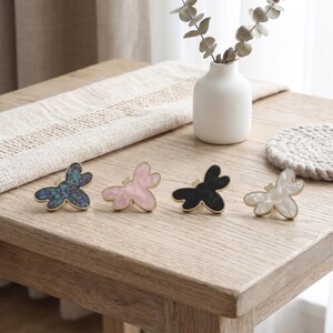 May include: Four butterfly-shaped hair clips with gold-toned edges, arranged in a row. The clips feature distinct finishes: one with a dark iridescent pattern, one in solid light pink, one in solid black, and one in a pearlescent white finish.