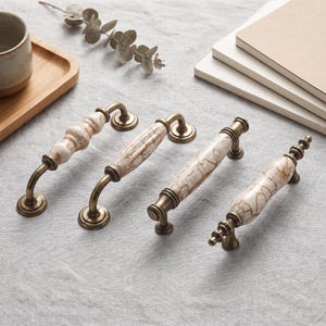 May include: Four cabinet pulls arranged in a row, each featuring a central ceramic handle with a cream and brown marbled pattern. The handles are supported by antique brass-toned metal hardware with circular mounting bases at each end.