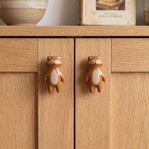 May include: Two brown and cream ceramic cabinet knobs shaped like stylized raccoons. Each knob features a smooth, glossy finish with a light-colored belly, pointed ears, and a small face with dark eyes, mounted on light-toned wooden cabinet doors.
