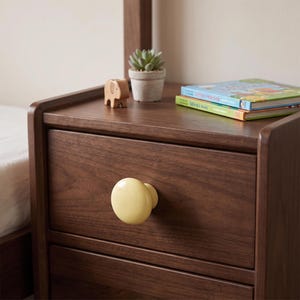 May include: A dark brown wooden nightstand featuring a single drawer with a smooth, round, pale yellow knob. On the top surface sits a small potted succulent, a wooden elephant figurine, and two stacked children's books with colorful covers.