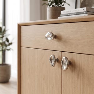 May include: A light oak wood cabinet featuring three irregular, organic-shaped silver metal knobs. One knob is centred on the top drawer, while two matching knobs are positioned on the lower cabinet doors, creating a reflective, metallic contrast.