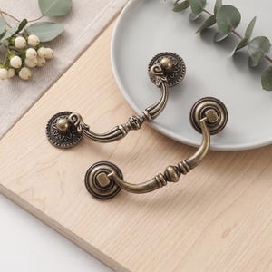 May include: Two antique brass-toned metal drawer pulls featuring a curved handle design with ornate, circular mounting plates. The hardware displays a weathered, metallic finish with intricate detailing on the circular bases and the central handle.