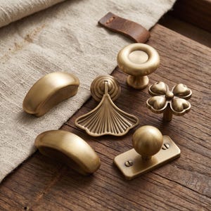 May include: A collection of six brass-toned metal cabinet hardware pieces, including two smooth arched pulls, a circular knob, a four-leaf clover shaped knob, a fan-shaped drop pull, and a spherical knob mounted on a rectangular base plate.
