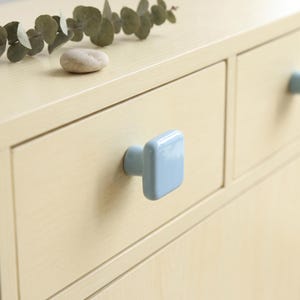 May include: A light blue square cabinet knob with a glossy finish attached to a light-colored wooden drawer. The knob features a smooth, rounded-edge square face and a cylindrical base, providing a simple and functional hardware component.