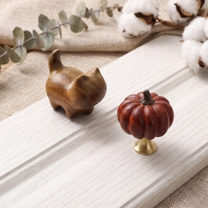 May include: A carved wooden cat figurine with a smooth brown finish and a small, round, reddish-brown wooden pumpkin knob featuring a metallic gold-toned base, both positioned on a white-painted wooden surface with visible wood grain texture.